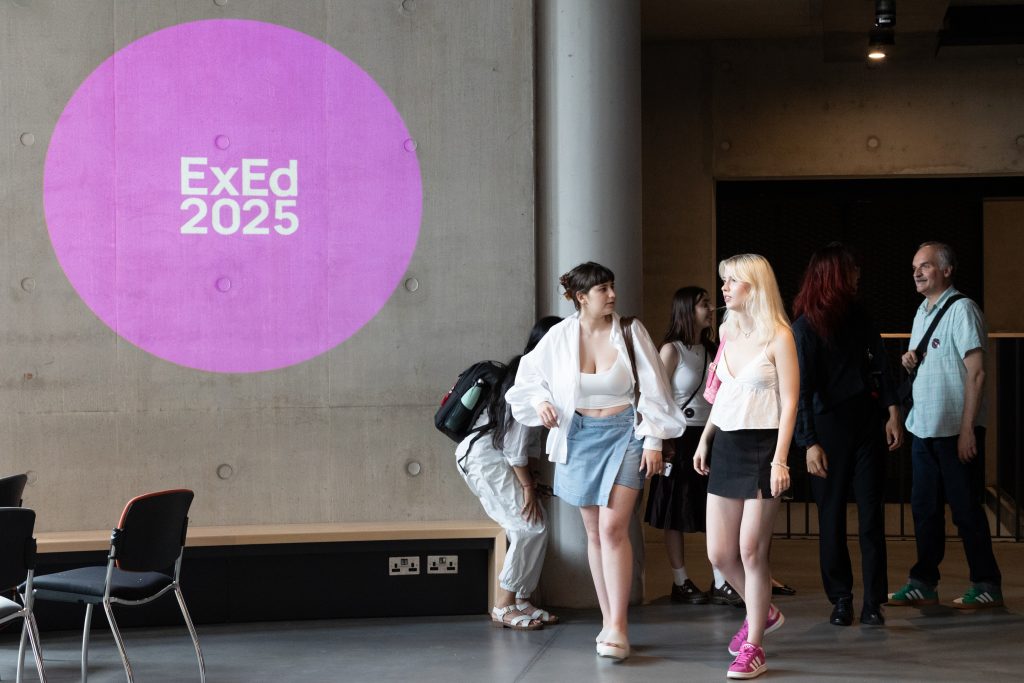 Participants walking by a concrete wall with a pink projection circle that reads ExEd25.